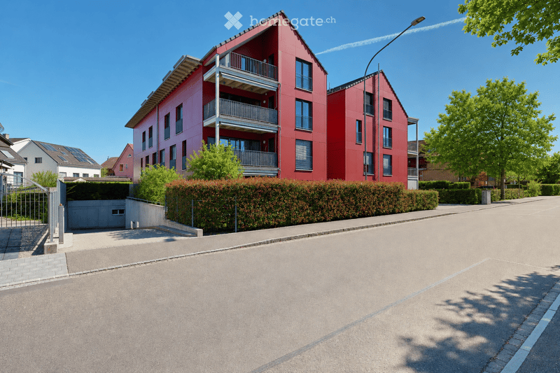 A modern red apartment building with balconies, surrounded by greenery and a paved driveway.