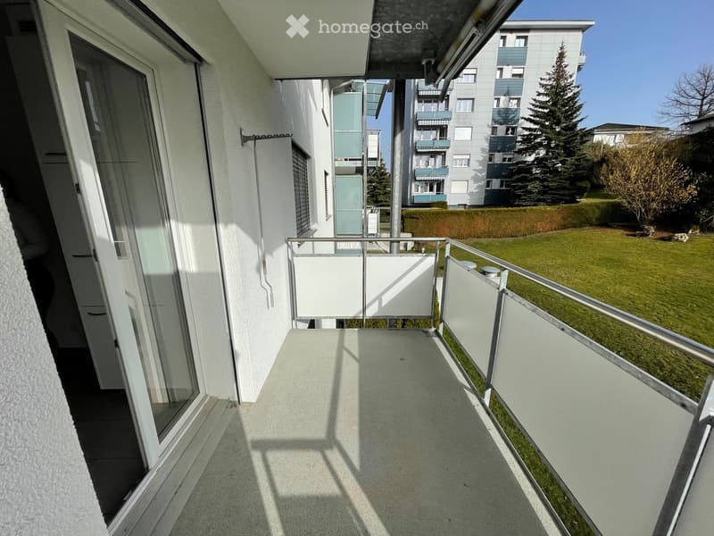 Balcony with modern glass railing, tiled floor, and view of a green lawn and neighboring buildings.