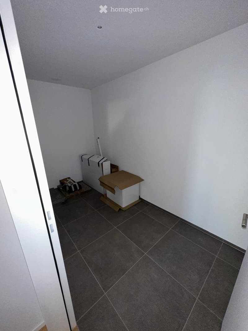 Empty room with white walls, dark tiled floor, and a partially open door on the left side. There are no visible furnishings or decorations.