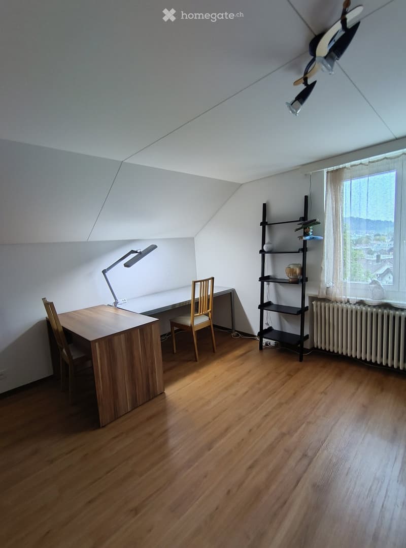 Bright, wooden-floored room with a slanted ceiling, featuring a wooden desk, chair, desk lamp, and a black shelving unit with decorative items.