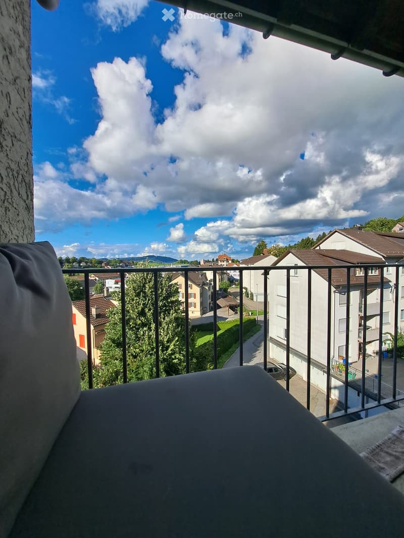 Balcony with metal railing, overlooking a residential area with houses, trees, and a clear blue sky with scattered clouds.