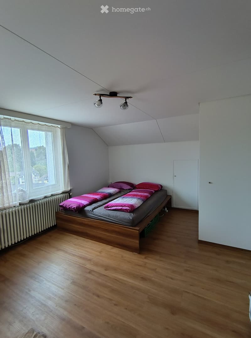 Bedroom with wooden floor, bed with pink and grey bedding, radiator, and large window with white curtains.