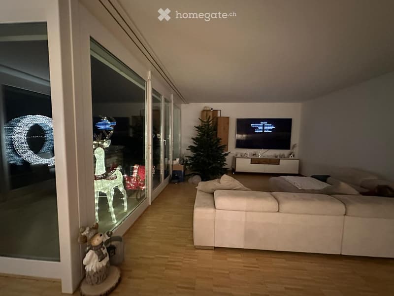 Modern living room with a white sectional sofa, flat-screen TV on a white stand, wooden floor, large glass sliding doors leading to a balcony, and a small Christmas tree in the corner.