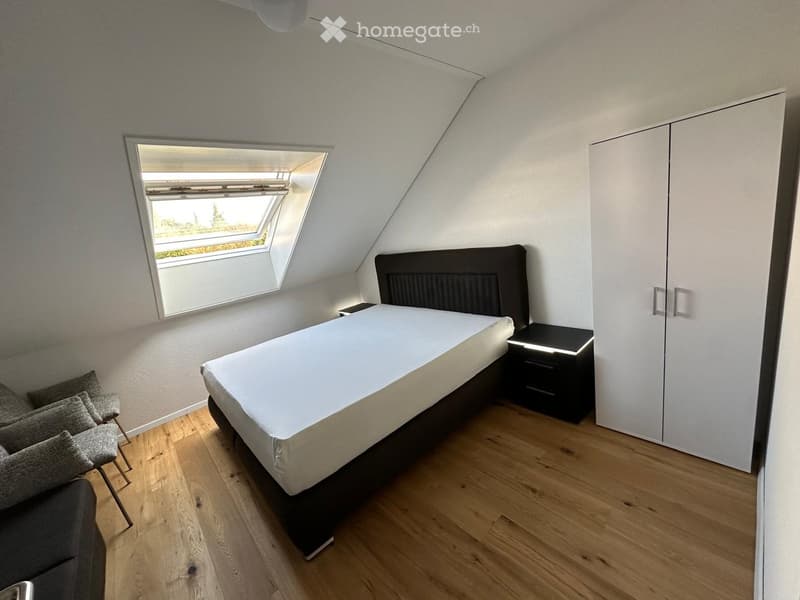 Cozy attic bedroom with wooden flooring, a single bed, nightstand, wardrobe, and a small seating area with chairs near a skylight window.