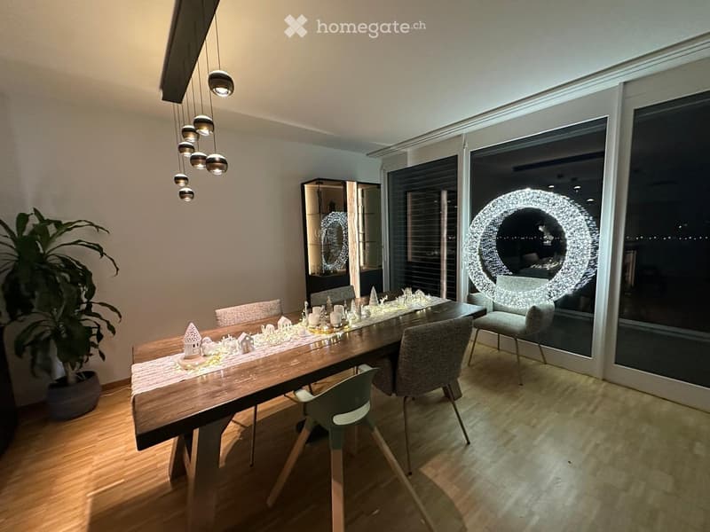 Modern dining room with a long wooden dining table, six chairs, a potted plant, and a mirror. The table is set with a table runner, candles, and decorative items.