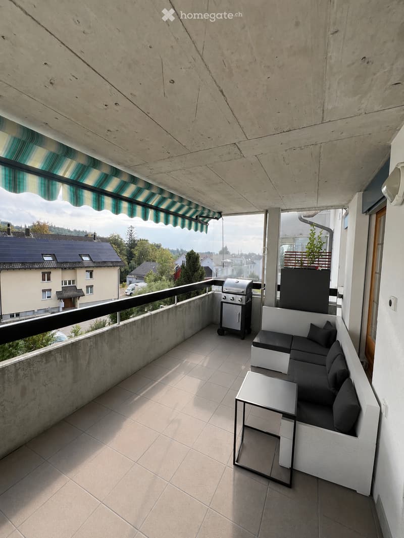 Modern balcony with a white sectional sofa, a small table, and a grill, featuring a striped awning and a view of the neighborhood.