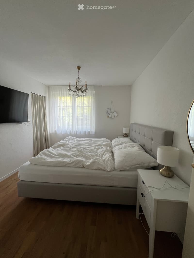 Modern bedroom with a large bed, nightstand, lamp, mirror, chandelier, and television mounted on the wall. The room features wooden flooring and large windows with white curtains.