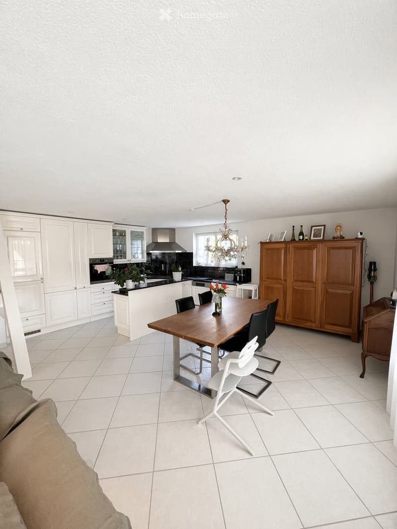 Bright, modern kitchen and dining area with white tiled flooring, white cabinets, a wooden dining table with chairs, and a chandelier hanging from the ceiling.