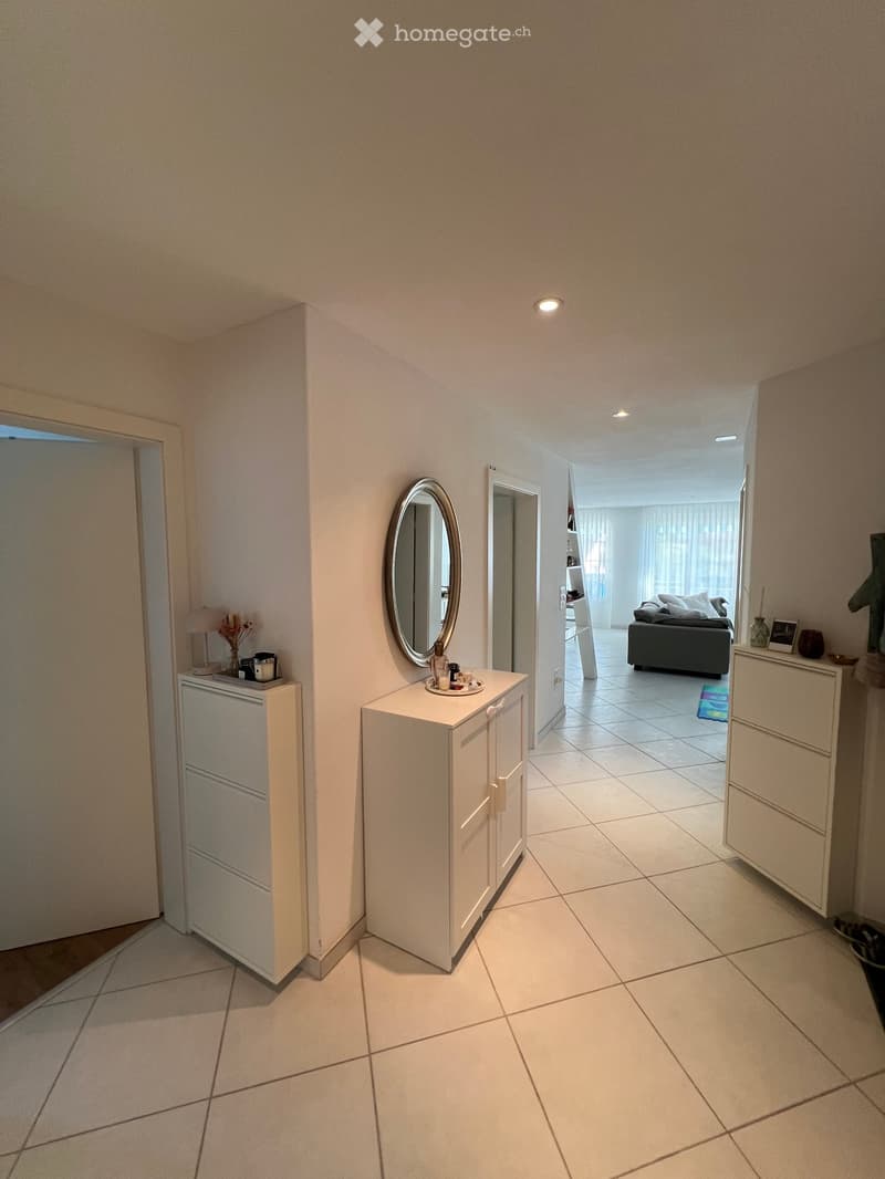 Hallway with tiled floor, white walls, small cabinets, and a mirror. A room with a sofa and decorative items is visible at the end.