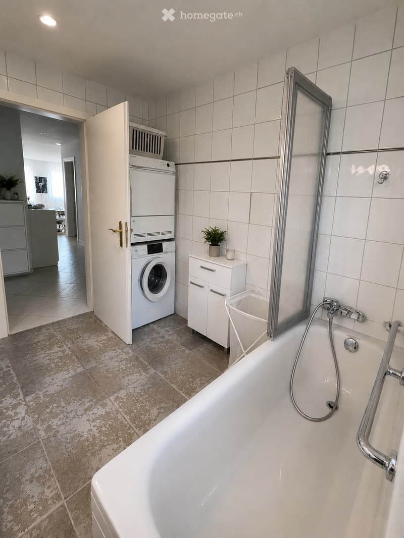Modern bathroom with a bathtub, white tiled walls, a washing machine, a small cabinet, and a potted plant on the counter.