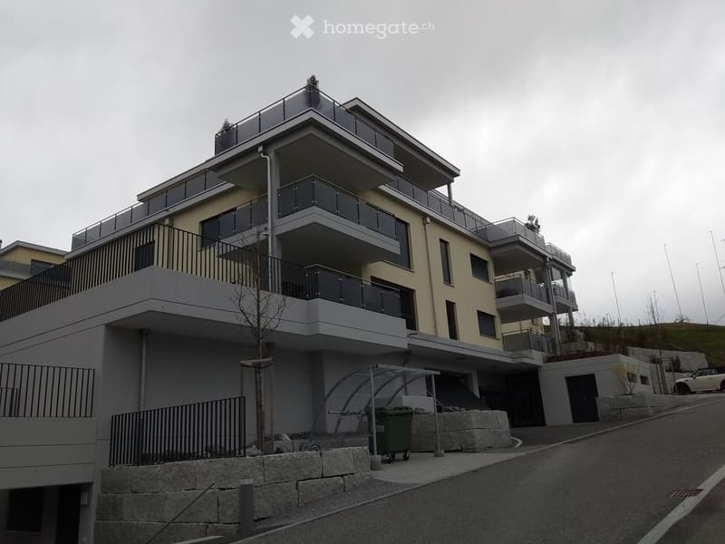 Modern multi-story residential building with balconies, glass railings, and a covered parking area.