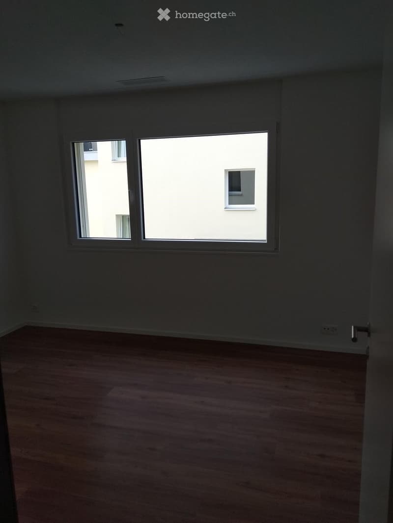 Empty room with wooden flooring and large windows, showing natural light and a view of neighboring buildings.