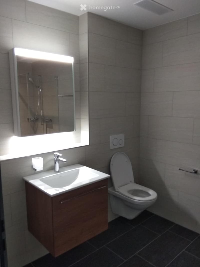 Modern bathroom with a wooden vanity, white sink, chrome faucet, toilet, large mirror with built-in lighting, and a small candle on the counter.
