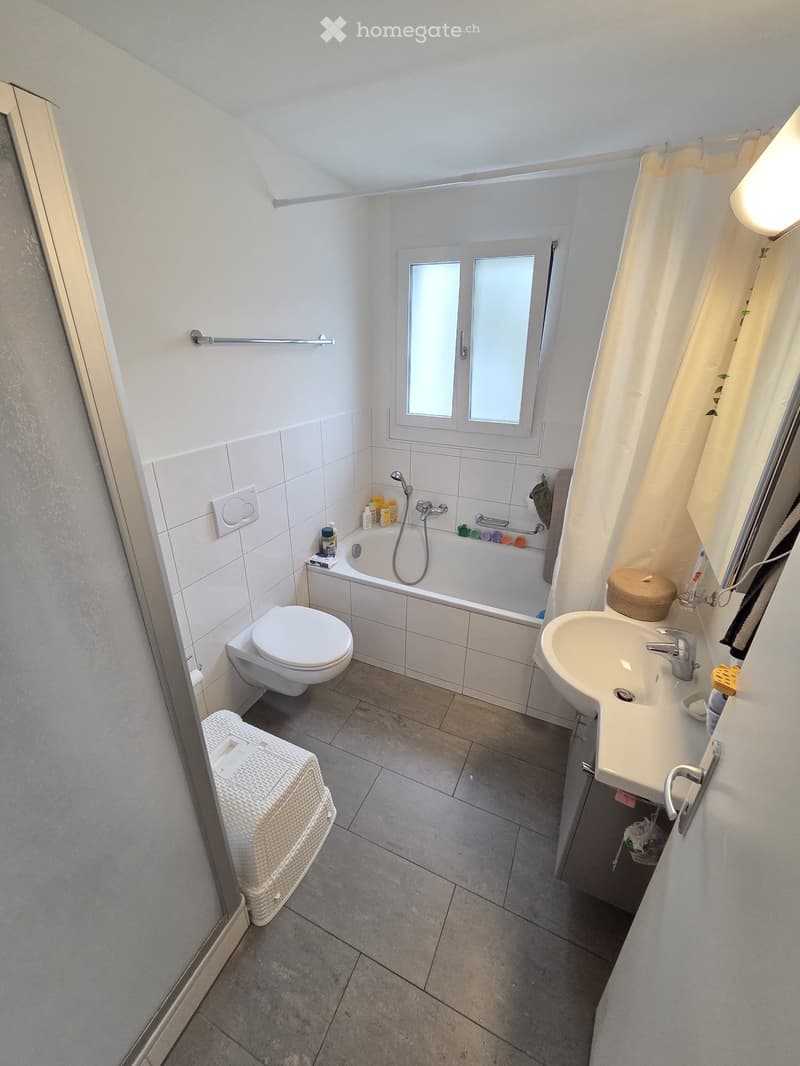 Modern bathroom with white tiles, a bathtub, toilet, sink, and a window with frosted glass. Includes a wicker basket and various toiletries.