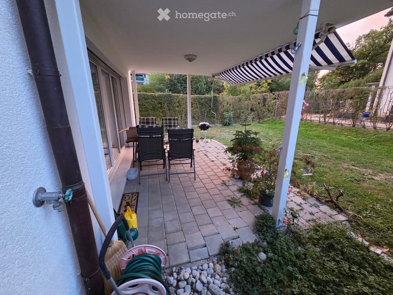 Balcony with two chairs and a small table, leading to a garden area with a grill and potted plants.