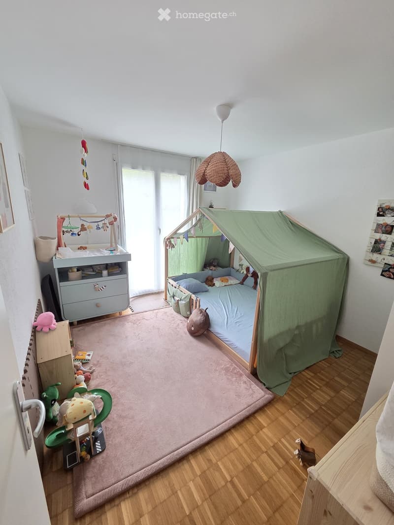 A well-lit children's bedroom with a wooden floor, a bed with a green tent, a crib, and various toys scattered around.