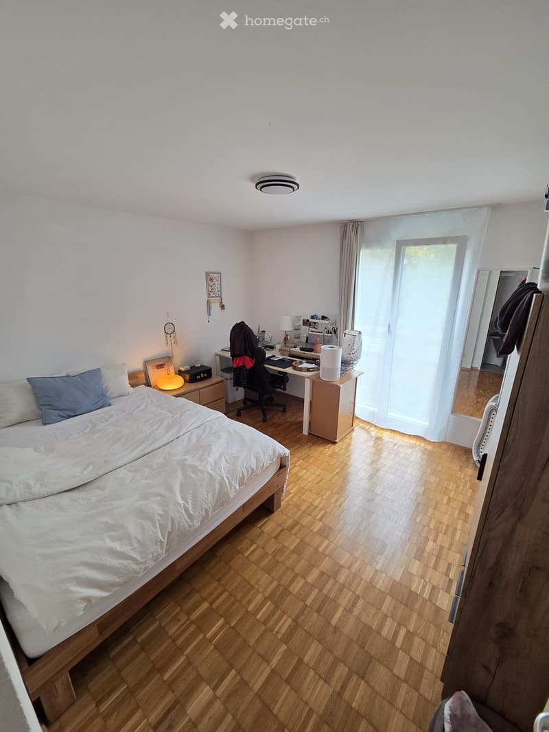 Cozy bedroom with a wooden bed, desk, chair, and nightstand. Features a large window with sheer curtains and a modern lamp on the nightstand.