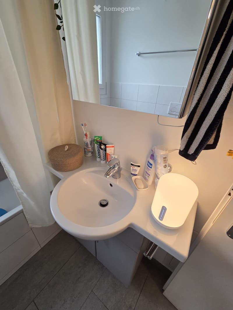 Modern bathroom with a white sink, chrome faucet, mirror, and various toiletries including a wicker basket, toothbrush holder, and soap dispenser. A towel hangs on a rack.