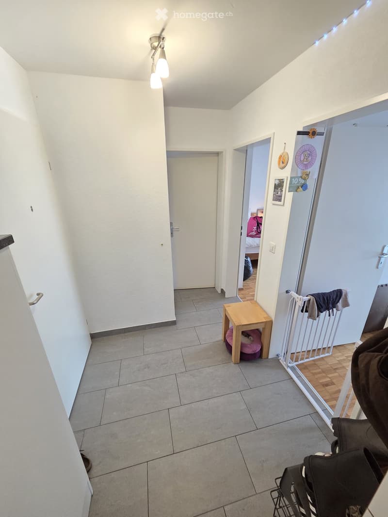A narrow hallway with tiled flooring, white walls, and a small wooden table with a pet bed. A baby gate and a stroller are visible on the right side.