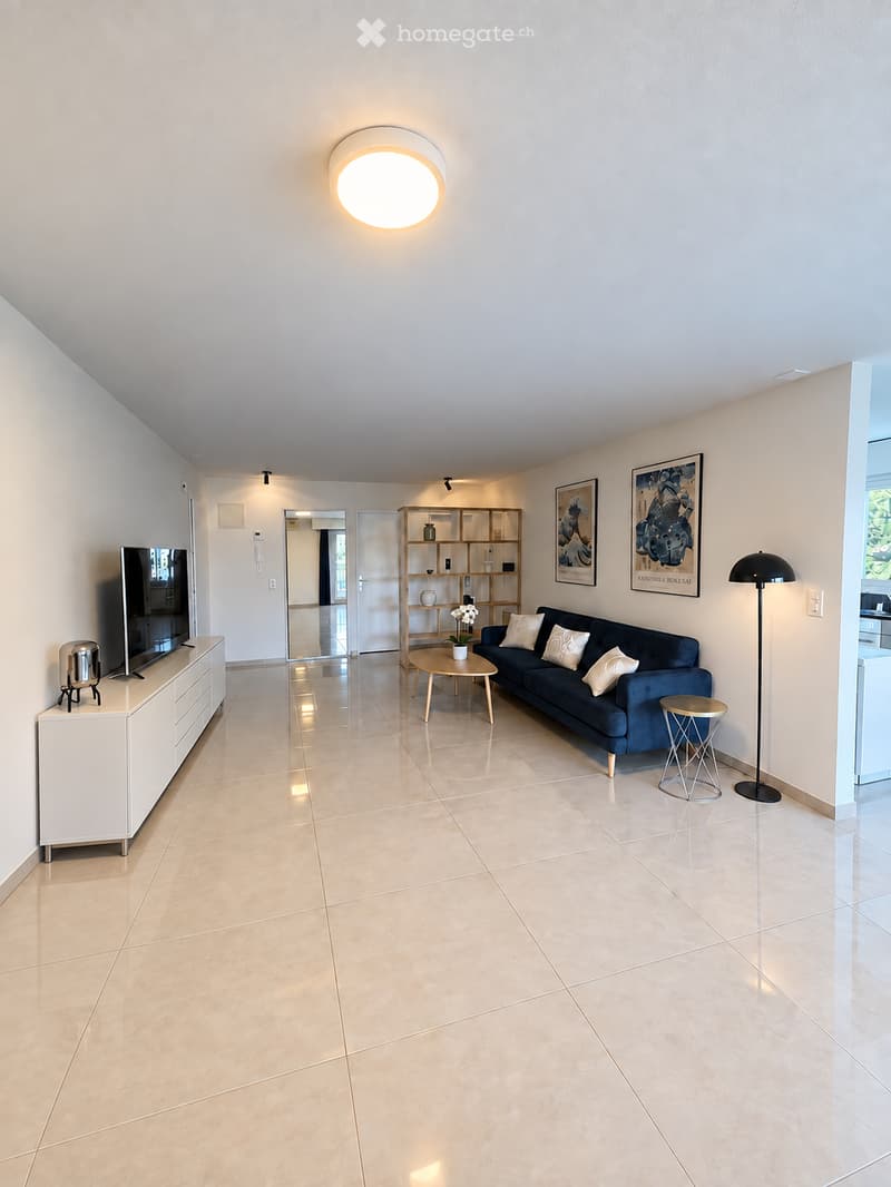 Modern living room with beige tiled flooring, a dark blue sofa with white throw pillows, a wooden coffee table, a white TV stand, a floor lamp, a small round stool, and a wooden shelf with decorative items.