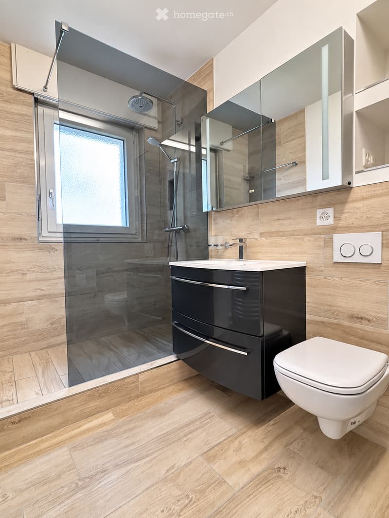 Modern bathroom with wooden walls and flooring, featuring a black vanity cabinet, white sink, toilet, large mirror, and a glass-enclosed shower with a modern showerhead.