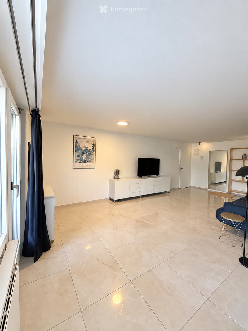 Modern living room with light-colored tiles, white walls, and large windows. Features a television on a white cabinet, a blue sofa, a small round table, and a floor lamp.