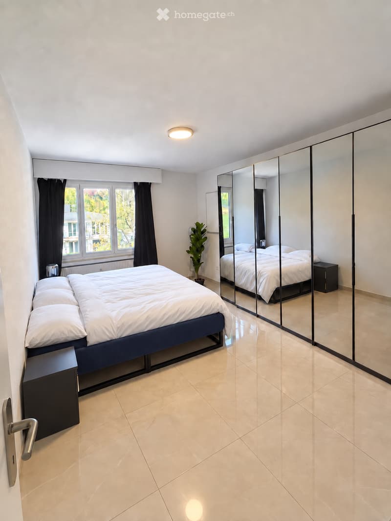 Modern bedroom with a large bed, nightstand, mirrored wardrobe, and a potted plant near the window.