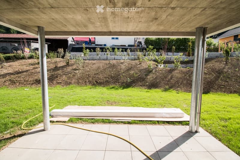 A modern outdoor patio with a wooden pergola, tiled floor, and a raised garden bed with young plants. The area is surrounded by a grassy lawn and includes a hose on the ground.