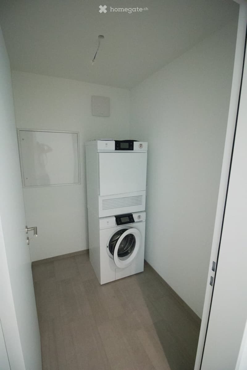 Small room with a white washing machine and dryer unit, light grey walls, and a wooden floor.