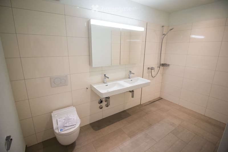 Modern bathroom with beige tiles, double sink, large mirror with built-in lighting, toilet, and shower area.