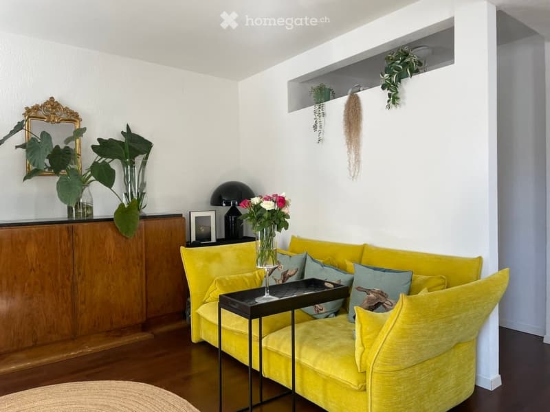 Bright living room with a yellow velvet sofa, coffee table, decorative mirror, vase with flowers, and potted plants on a wooden cabinet.