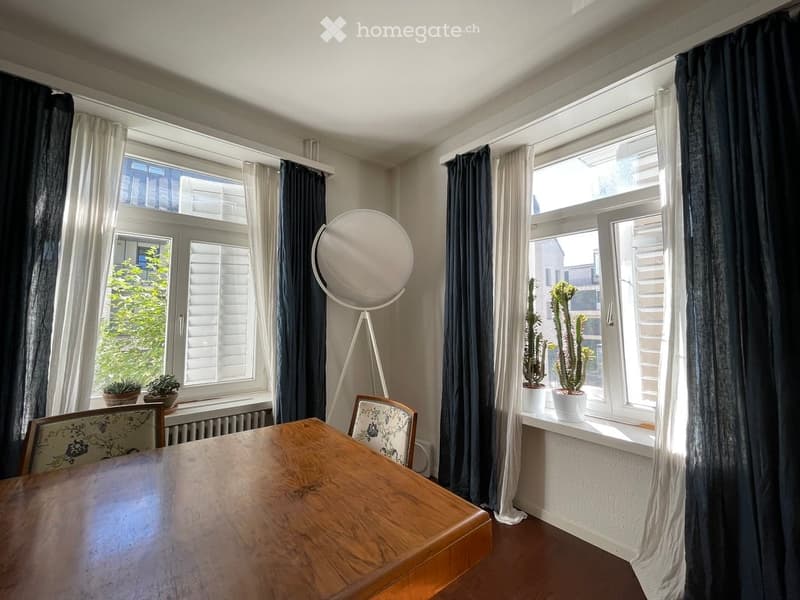 Bright room with large windows, wooden table, chairs, floor lamp, and potted plants on the windowsill.