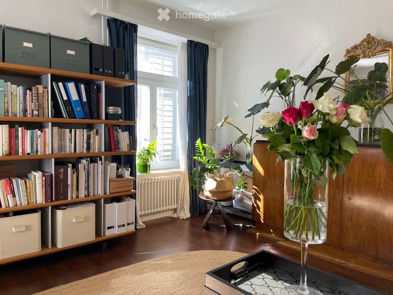 A cozy, well-lit room with wooden flooring, large window with dark curtains, bookshelf filled with books and storage boxes, small wooden stool with a woven basket, vase with flowers, and a decorative mirror on the wall.