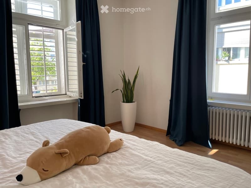 Cozy bedroom with a large stuffed animal on the bed, a potted plant, and large windows with dark curtains.