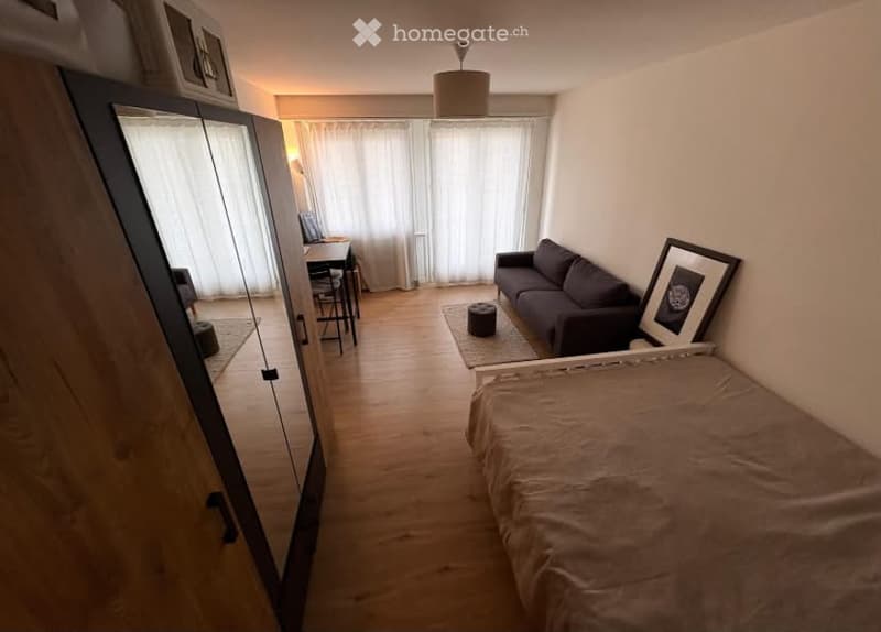 A compact, well-lit room with a bed, sofa, wardrobe, and a small table with chairs. The space features wooden flooring and large windows with white curtains.