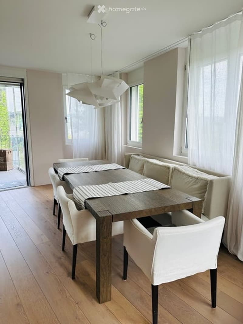 Dining area with a wooden table, white chairs, and a sofa, large windows with white curtains, and a modern chandelier.