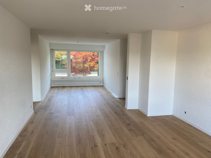 Empty room with light wooden flooring, white walls, and a large window with a view of autumn foliage.