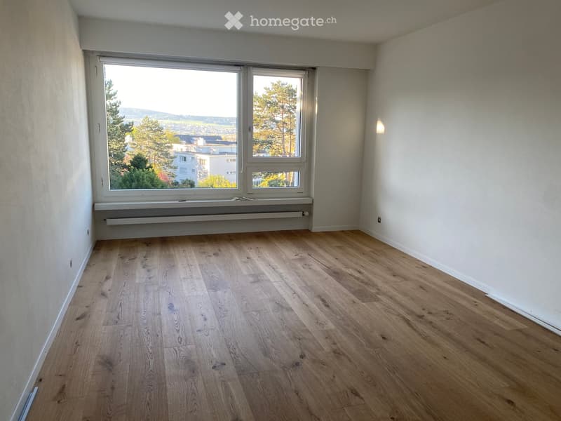 Empty room with wooden flooring, white walls, and large windows offering a scenic view of trees and buildings.