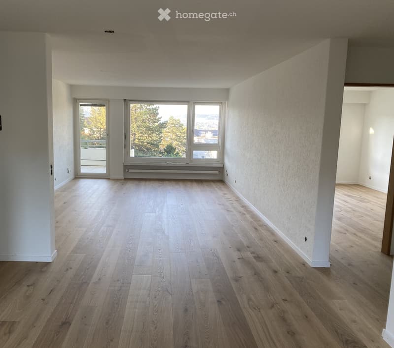 Empty room with light wooden flooring, white walls, large windows, and a sliding glass door leading to a balcony.