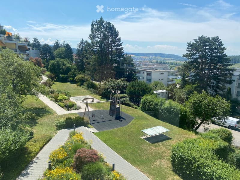 A well-maintained garden area with a swing set, picnic table, and pathways surrounded by trees and shrubs, with residential buildings in the background.