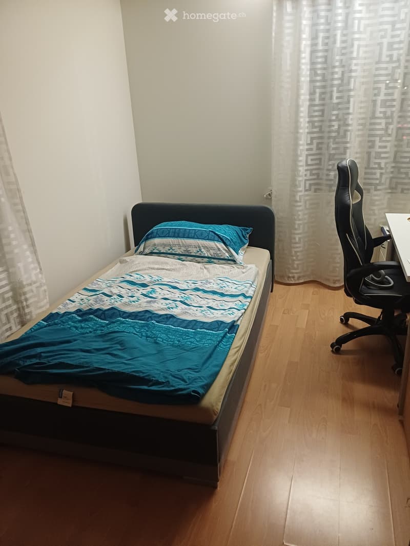 A small, furnished bedroom with a single bed, a desk, and a chair. The bed is made with blue and white bedding, and the room has wooden flooring and white walls.