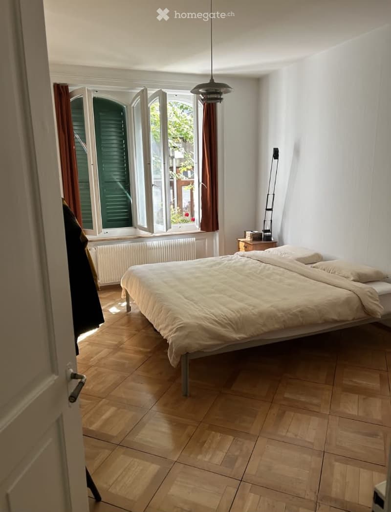 Bright bedroom with wooden flooring, large window with open shutters, bed with beige bedding, nightstand with lamp, and radiator below the window.