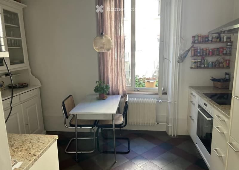 A compact kitchen with a small dining area. Features include a white table with two chairs, a potted plant, built-in cabinets, an oven, and shelves with various items.