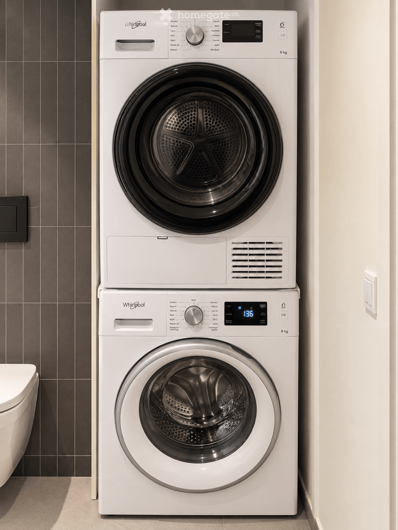Modern bathroom with a white Whirlpool washing machine and dryer stacked vertically, a toilet, and dark tiled walls.