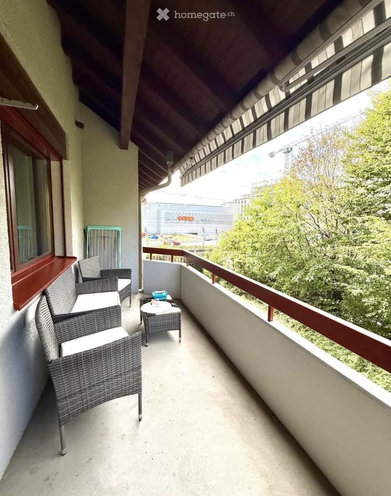 Balcony with wicker chairs, small table, and a view of greenery and a building.