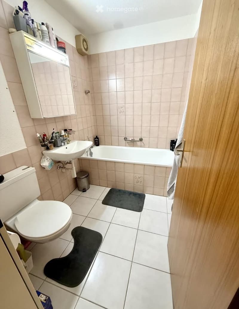 Bathroom with a toilet, sink, bathtub, mirror, and cabinet. The floor is tiled, and there are two black bathroom mats placed in front of the bathtub and toilet.