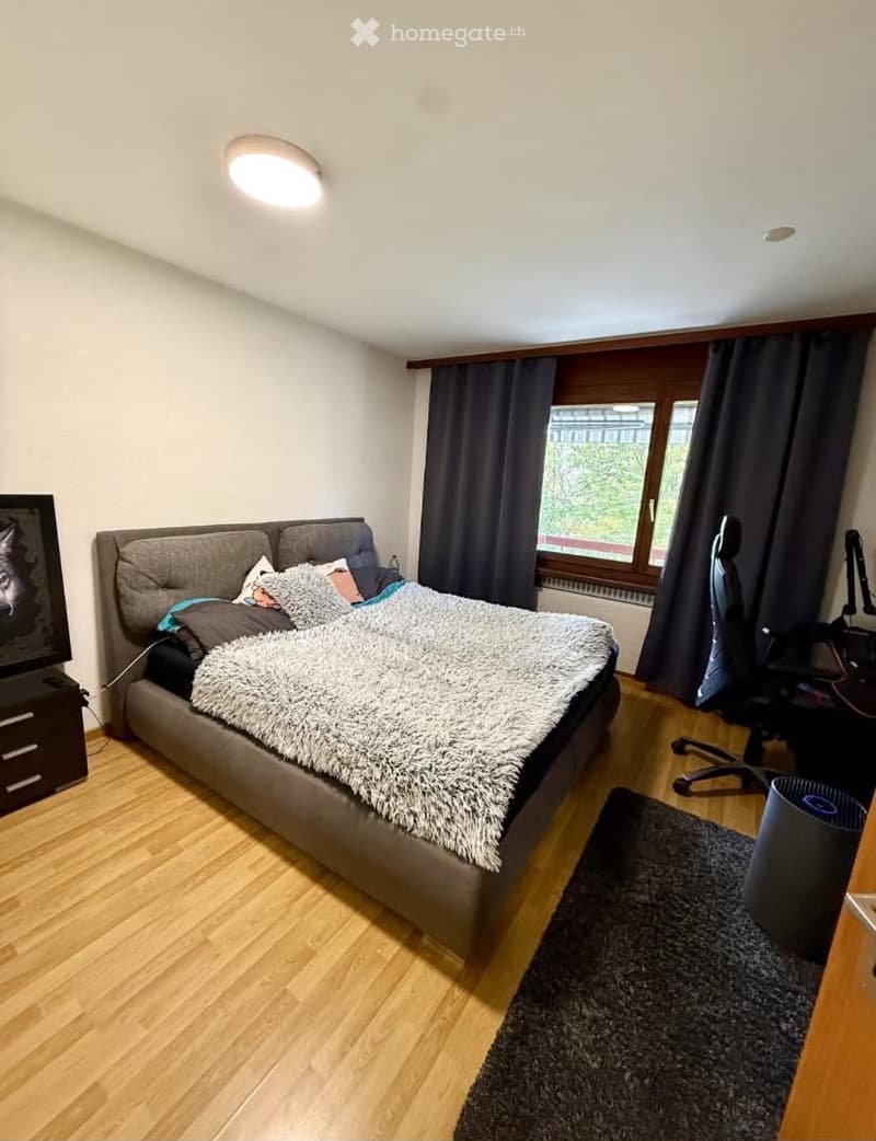 A modern bedroom with a large bed, a desk, a chair, and a TV screen. The room features wooden flooring, large windows with dark curtains, and a cozy, well-lit atmosphere.