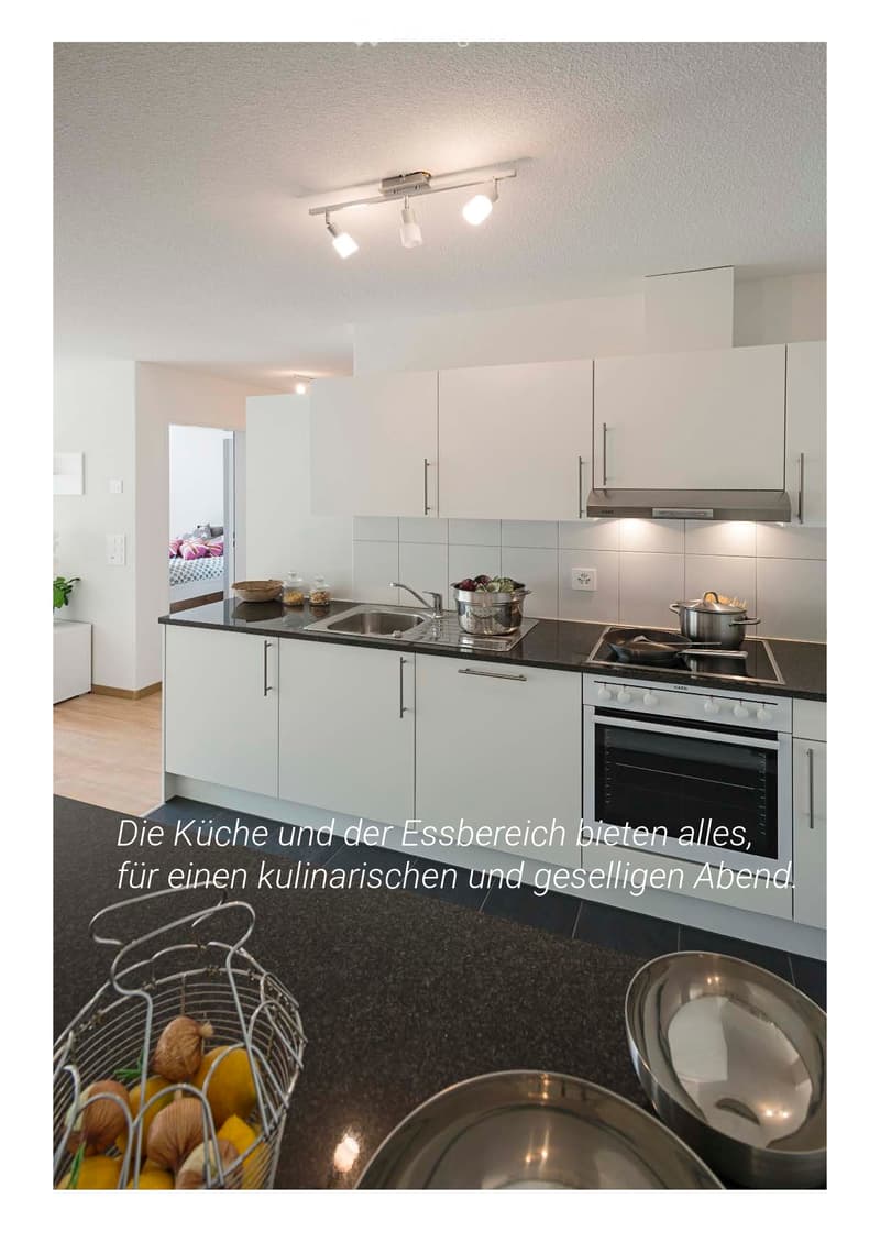 Modern kitchen with white cabinets, black countertops, stainless steel appliances, and a sink. Visible items include a pot, pan, bowl, and a basket with fruits.
