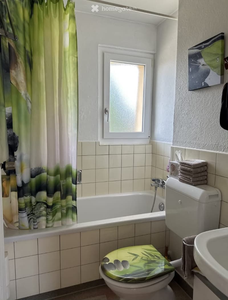 Bathroom with a bathtub, toilet, sink, and a green bamboo-patterned toilet seat cover. The room features tiled walls and a window with a curtain.