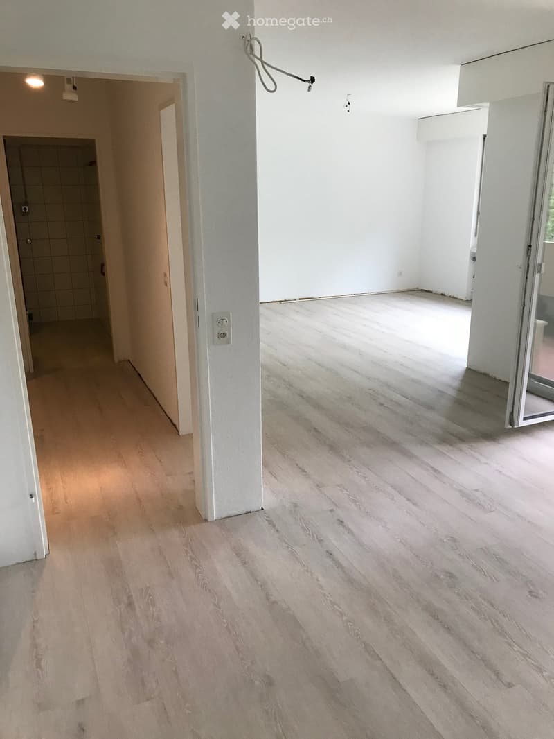 Empty hallway with light wooden flooring and white walls, leading to a tiled bathroom area.
